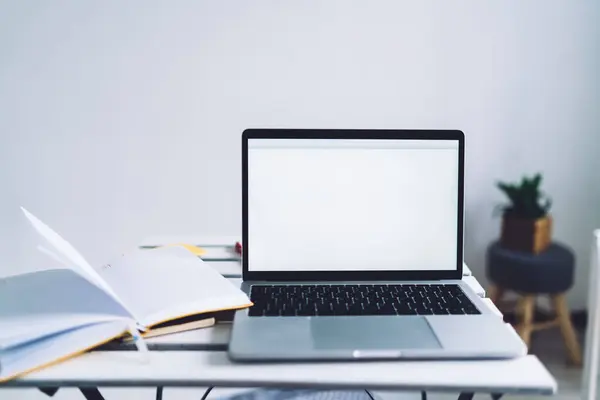 明亮房间里的工作空间 有白色的木制桌子 上面放着灰色的笔记本电脑 屏幕是白色的 空白页上有打开的笔记本 — 图库照片