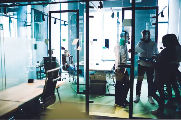 Resmi olarak kadın ve erkek çalışanlar, modern ofiste cam arkasında şirket stratejisini tartışan belgeler taşıyorlar.