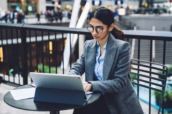Gri ceketli ve gözlüklü kendine güvenen etnik iş kadını New York 'ta, sokak terasında masada otururken tablet üzerinde çalışıyor.