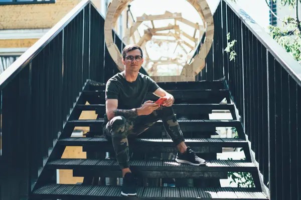 Genç, sıska, gözlüklü, haki renkli gözlüklü, güneşli yaz gününde merdivenlerde oturmuş, cep telefonuyla sörf yapan bir erkek.