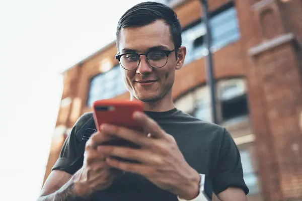 Below view of millennial smiling hipster guy playing online games via application on cellular phone while waiting friend on urban setting and using public internet connection, concept of generation