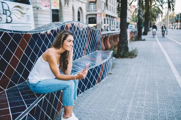 Side view of beautiful positive smiling female in stylish outfit surfing smartphone during chilling on decorative city bench looking away