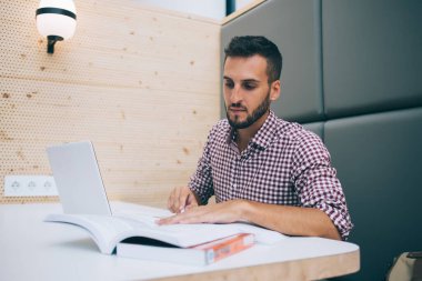 Gündelik giysiler içinde odaklanmış genç bir adam kafeteryada oturmuş bilgi arayan ders kitabı okumaya hazırlanıyor.