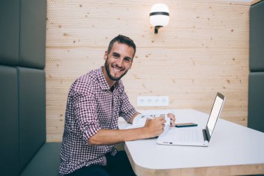 Keyifli gülümseyen serbest çalışan iş yerindeki masada oturmuş kameraya bakarak dizüstü bilgisayar kullanarak proje üzerinde çalışan biri.