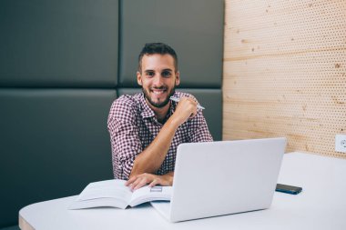 Keyifli serbest çalışan, elinde kalemle çalışan, dizüstü bilgisayar okuyan, kitap yazan, modern kafede oturmuş kameraya bakan biri.