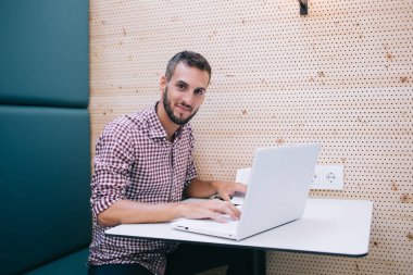 Keyifli gülümseyen, sakallı, normal tişörtlü, mavi koltukta oturan, duvarında lamba olan bir erkek.