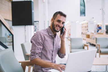 Dizüstü bilgisayarla çalışan ve yaratıcı iş yerinde cep telefonuyla konuşan sıradan, mutlu, sakallı, genç bir erkek.