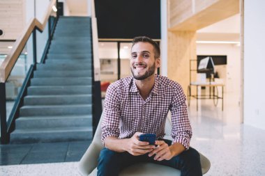 Neşeli, güler yüzlü, sakallı, normal tişörtlü, kot pantolonlu, yaratıcı iş yerinde sandalyede oturan, cep telefonuyla mesajlaşan bir erkek.