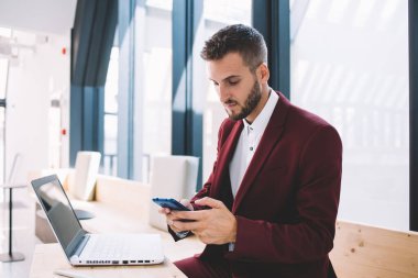 Klasik takım elbiseli, konsantre yönetici sakallı bir adam cep telefonuyla mesajlaşırken aynı anda iş yerinde dizüstü bilgisayarın önünde oturuyor.