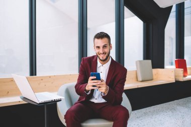 Beyaz gömlekli ve bordo takım elbiseli, kendine güvenen bir erkek bilgisayarın yanında oturmuş kameraya bakarken cep telefonunu karıştırıyor.