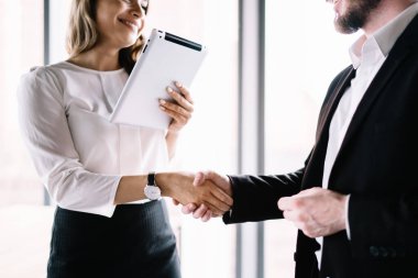 Tabletle gülümseyen kadın ofis çalışanı ve resmi kıyafetlerle siyah takım elbiseli meçhul erkek girişimciyle el sıkışıyor.