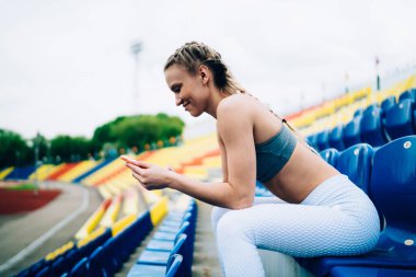 Spor kıyafetleri içindeki neşeli sarışın bayan sporcunun stadyum koltuğunda bulanık arka planda otururken akıllı telefonuyla mesajlaşması.
