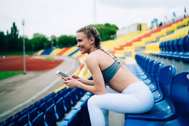 Spor kıyafetleri içindeki neşeli sarışın bayan sporcunun akıllı telefonla mesajlaşması ve stadyum koltuğunda otururken ileriye bakması.