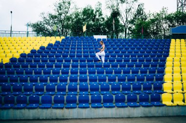 Sıska kadın spor kıyafetleri içinde şehir stadyumunun renkli tribünlerinde duruyor ve yeşil ağaçlara karşı sörf yapıyor. 