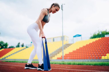 Beyaz taytlı zayıf bir kadın mavi lastik kurdeleye basıyor ve arka planda özenle antrenman yapıyor.