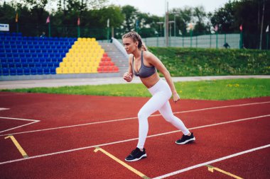 Beyaz taytlı, gri tişörtlü, koşan pistte duran ve stadyuma koşmadan önce yüzünü çeviren bir kadın.