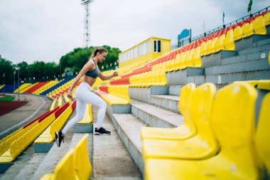 Spor odaklı, at kuyruklu, merdivenlerde bacak egzersizi yapan ve stadyumdaki tribünlere yoğunlaşan kadınların yan görünüşü.