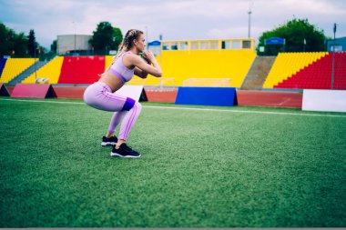 Spor kıyafetleri içinde spor giyinen genç bir kadının yeşil çimlere çömelmiş lastik bantla spor yaparken şehir stadyumunun tribünlerine baktığı yan manzara. 