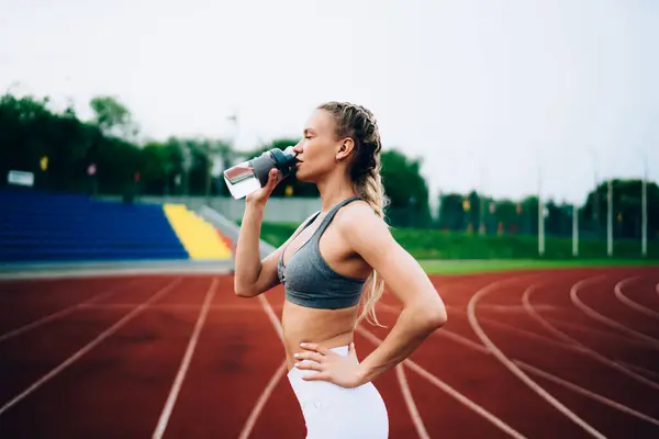 スタジアムのジョギングコースの背景に立っている間 スポーツボトルから水を飲む若いフィットの女性アスリートの側面のビュー — ストック写真
