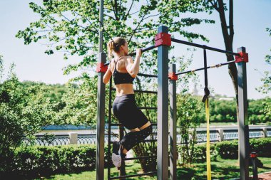 Açık renk saçlı bayan sporcu halter üzerinde barfiks egzersizi yapıyor ve güneşli bir günde, göl kıyısındaki parkın yanındaki koşu pistinin yanındaki ekipmanları kullanıyor.