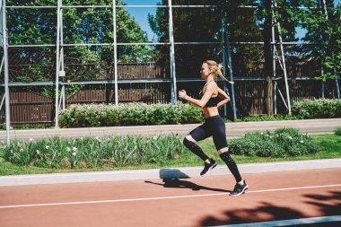 Genç atletik sarışın bayan koşucunun yan görünüşü ve güneş ışığında spor sahasında koşmaya devam etmesi.