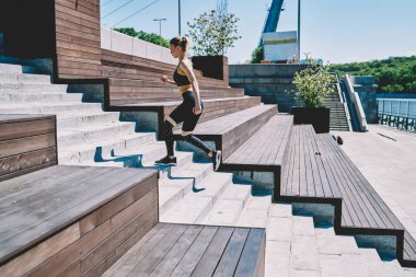 Spor kıyafetleri içinde, parlak güneşin altında rıhtımdaki merdivenlerde egzersiz yapan konsantre genç bir kadının yan görüntüsü.
