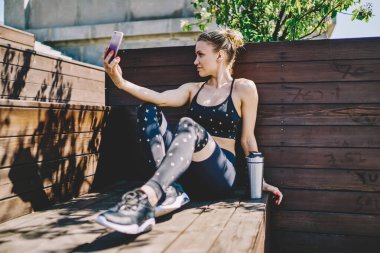 Şık giyinen modern genç sporcu kadın fiziksel eğitim sırasında kentteki ahşap bankta dinlenirken akıllı telefondan selfie çekiyor. 