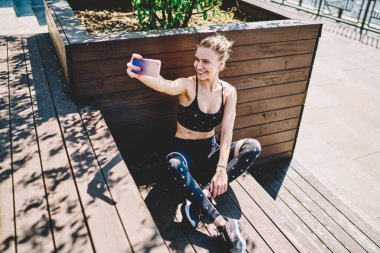 Havalı, sarı saçlı, şık, aktif giyinen, ayakları çapraz oturan ve parkta cep telefonuyla selfie çekerken gülümseyen neşeli, sportif kadından. 