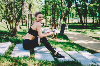 Spor kıyafetli, kulaklıklı, mutlu, gülümseyen bir kadın. Cep telefonu takıyor, egzersiz yapıyor, merdivenlerde kameraya bakıyor.