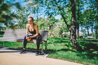 Siyah bluzlu ve taytlı neşeli enerjik bayan parkta elinde termos şişesiyle bankta yeşil ağaçlar ve çalılarla oturuyor.