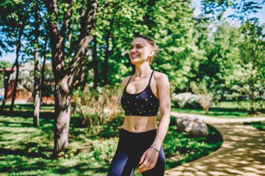 Enerjik, güler yüzlü, zayıf, sportif bir bayan. Siyah bluz ve tayt giymiş. Yazın arka planda yeşil ağaçlar ve çalılarla park yolunda yürüyor.