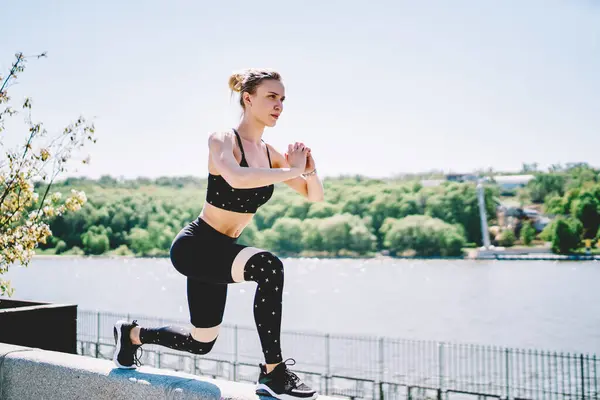 Concentrated sportive woman exercising and crouching bending leg and folding hands before chest on concrete fence along river in bright day