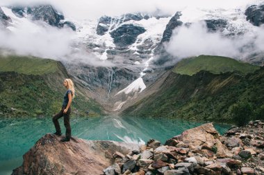 Aktif giyinmiş kadın yürüyüşçü, kar bulutlarıyla kaplı dağlarda gezinme arzusu sırasında tepedeki kayanın tepesinde durarak giyiniyor. Turist, pikoresk platolarla doğal çevrenin güzelliğinin tadını çıkarıyor.