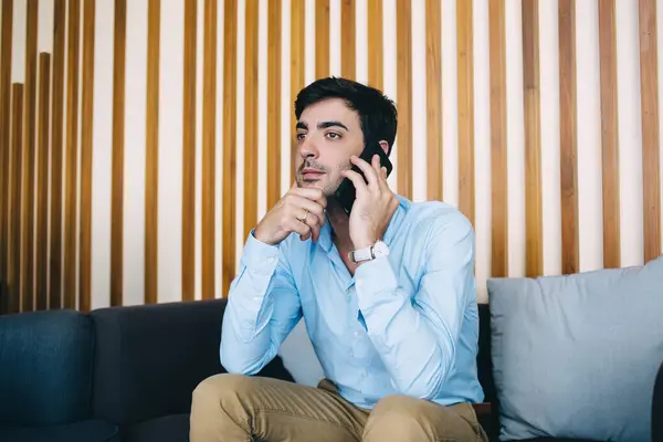 Pensive spanish businessman in formal outfit and luxury lifestyle pondering while talking on mobile phone about work, young proud serious male ceo banking via smartphone conversation indoors