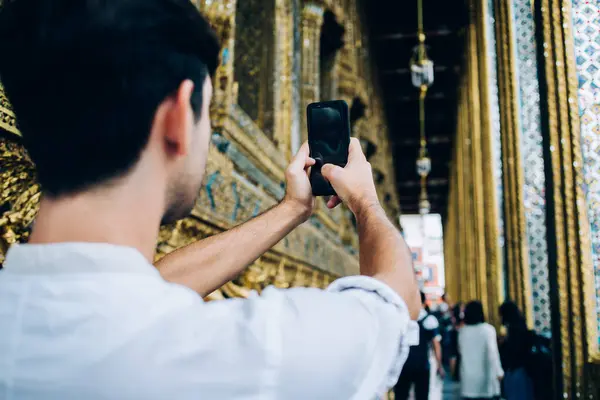 Back view of man holding mobile phone shooting video on mobile phone visiting golden asian temple, male traveler using smartphone camera taking photo for share in travelblog during journe