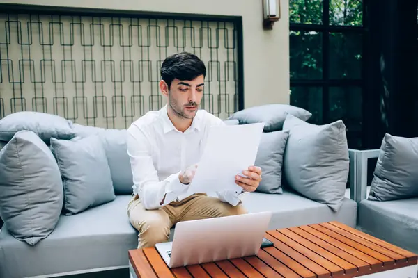 Thoughtful concentrated male entrepreneur reading document with data while using technology on remote job, confident businessman sitting next to laptop computer analyzing online income