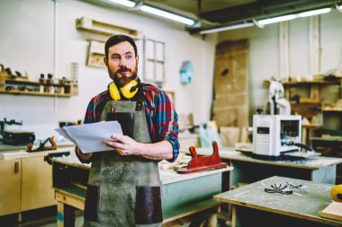 Önlüklü düşünceli bir genç adam kendi atölyesinde mobilya yapmaya hazırlanan projenin taslağını inceliyor.
