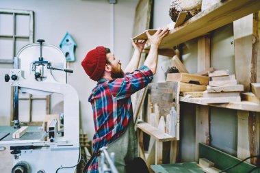 İş yerinde ahşap mobilya yapımına hazırlanmak için üst raftan malzeme alan önlüklü ve şapkalı adamın yan görünüşü