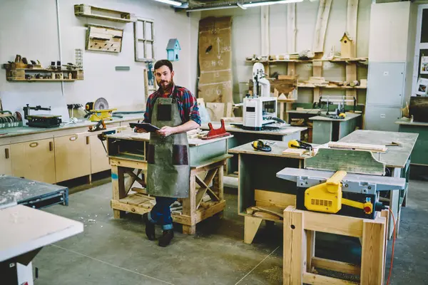 Full body positive bearded man in apron and with notepad leaning on workbench and looking at camera while working in shabby joinery