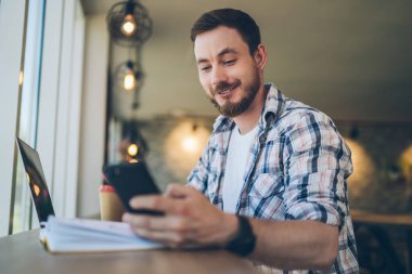 İyimser gülümseyen sakallı, kareli tişörtlü, akıllı telefonuyla sırıtan, iş projesini uzaktan takip eden, odadaki dizüstü bilgisayarlı adam.
