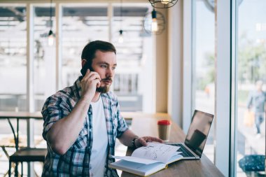 Genç, düşünceli, sıradan bir adam telefonda konuşuyor. Kafede çalışıyor. Dizüstü bilgisayarını tutuyor ve not defterini karıştırıyor.