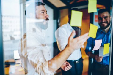 Zarif klasik gömlekli akıllı, çoklu etnik çeşitlilikte iş ortakları konuşuyor ve ofisteki pencerelerdeki sarı çıkartmalar hakkında bilgi topluyor.