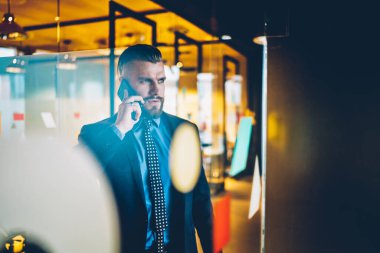 Camdan kendine güvenen, takım elbiseli, sakallı bir iş adamının modern ofis koridorunda dikilirken cep telefonuyla konuşması.