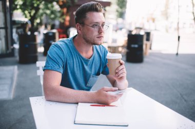 Düşünceli beyaz erkek blogcu gözlüklü terasta kahvesini yudumlarken sosyal ağlarda akıllı telefonla yayın yapma fikrini düşünüyor yaratıcı yazar açık havada geziniyor. 