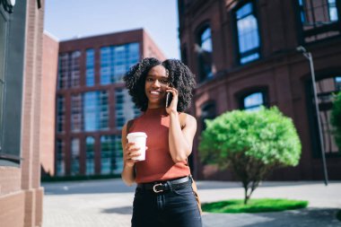 Neşeli etnik bayan, günlük giysiler içinde elinde kahve bardağıyla yazın şehirde cep telefonundan arama yapıyor. 