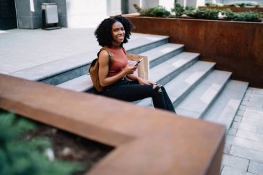 Afro-Amerikan kıvırcık saçlı, günlük kıyafetler giyen, akıllı telefon kullanan, sosyal ağları kontrol eden şehrin merdivenlerinde kameraya bakan neşeli Afro-Amerikan kadının yan görüntüsü.