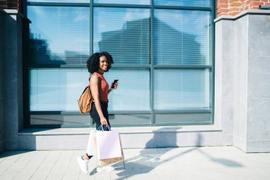 Neşeli, güler yüzlü, gündelik kıyafetli genç siyahi kadın yazın şehirde sokakta sırt çantasıyla geziyor.