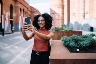 Afro saç stili, günlük kıyafet ve gözlüklü, telefon kullanarak selfie çeken güneşli bir günde sokakta gezen parlak siyah kadın.