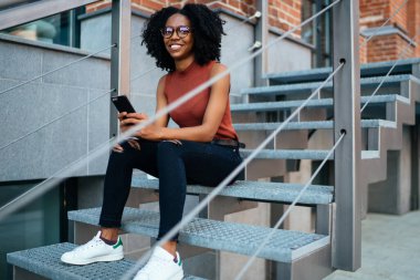 Siyah, nazik, Afro saçlı, günlük kıyafetleri ve gözlükleriyle gülümseyen bir kadın akıllı telefon kullanarak şehirde merdivenlerde oturmuş kameraya bakarak eğleniyor.