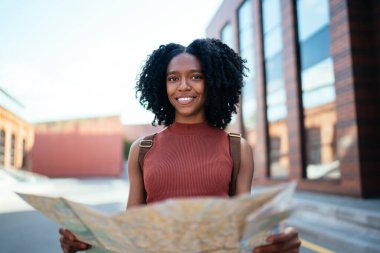Parlak, kıvırcık saçlı, sıradan giyinmiş, gülümseyen bir kadın. Ünlü şehrin sokaklarında dolaşıp kameralara bakıyor.
